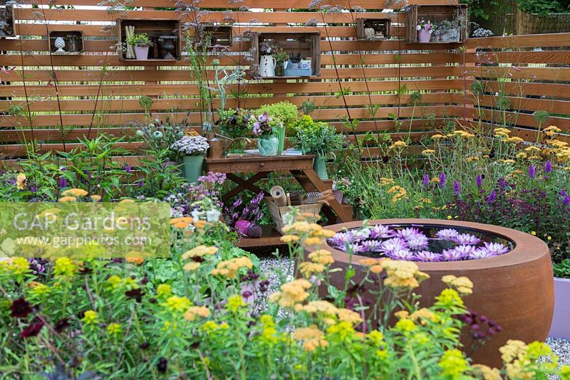 Urbis water feature surrounded by raised beds, crates used as shelves on a slatted fence - Katie's Lymphoedema Fund: Katie's Garden, RHS Hampton Court Palace Flower Show 2016