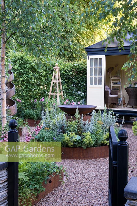 Gateway into garden with gravel path leading to a Summer House, round border with rust effect water bowl - CCLA: A Summer Retreat, Design: Amanda Waring and Laura Arison, RHS Hampton Court Palace Flower Show 2016.