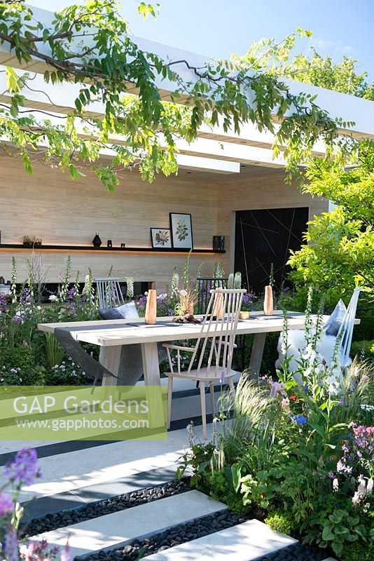 The LG Smart Garden, view of a patio with wooden chairs, table by Nils Verweij, pergola and the path made from limestone paving slabs and separated with black oval stones surrounded by Stipa tenuissima, Tiarella 'Spring symphony', Digitalis purpurea f. albiflora 'Alba', Wisteria. Designer: Hay Young Hwang, Sponsors: LG Electronics, RHS Chelsea Flower Show 2016
