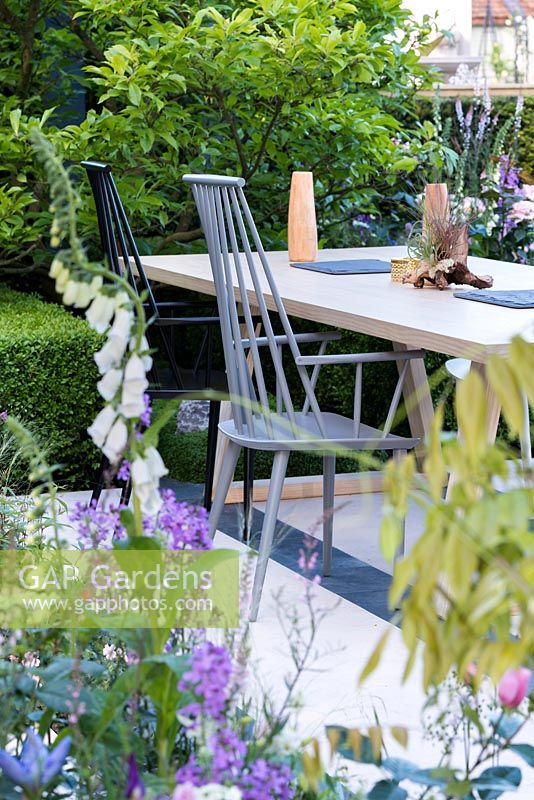 The LG Smart Garden, view of a patio with wooden chairs and table designed by Nils Verweij surrounded by spring flowers in pale colours. RHS Chelsea Flower Show 2016. Designer: Hay Young Hwang, Sponsors: LG Electronics