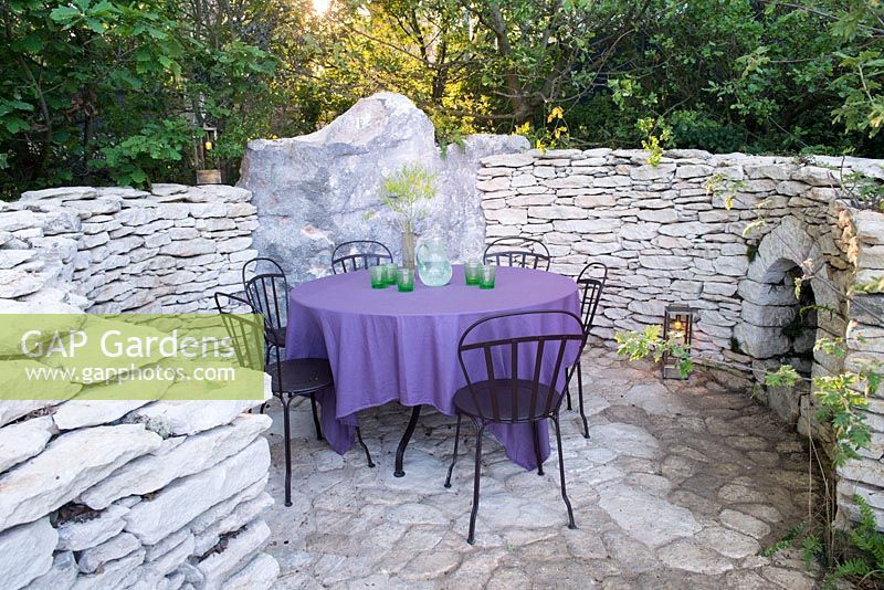 L'Occitan Garden, view a patio with black metal chairs, table with purple table cloth, glasses, jug and lanterns surrounded by curved sand stone fence with fire place.  RHS Chelsea Flower Show, 2016.