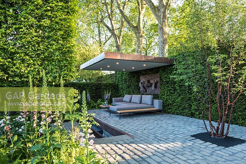The Husqvarna Garden, view of modern patio seating area with furniture, bronze sculpture, bluestone cobbled paths, surrounded by  Digitalis purpurea, Prunus maackii 'Amber Beauty', Carpinus pleached floating cubes, Taxus hedging. RHS Chelsea Flower Show, 2016.
