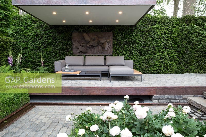 The Husqvarna Garden, view of outdoor living area on floating stone terrace with sofa and cushions, covered canopy with lighting, copper water rill, Taxus baccata hedge, Paeonia 'Elsa Sass' and Digitalis purpurea - Brass sculpture by Willy Wildlife Australia
