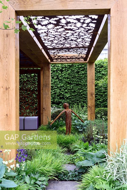 The Morgan Stanley garden for Great Ormond Street Hospital. Rusty metal stenciled screen above pergola walk in timber frame. Rusting metal figure of young boy in woodland garden. RHS Chelsea Flower Show 2016
