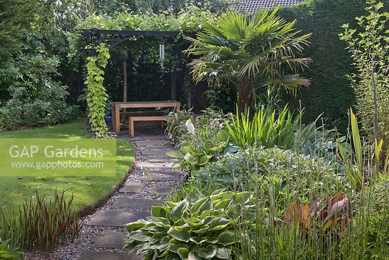 Indian stone path leading past tropical style flower bed and lawn to black stained wooden pergola with Humulus lupulus 'Aureus' and grape vine covering oak table and seating - July, Cheshire