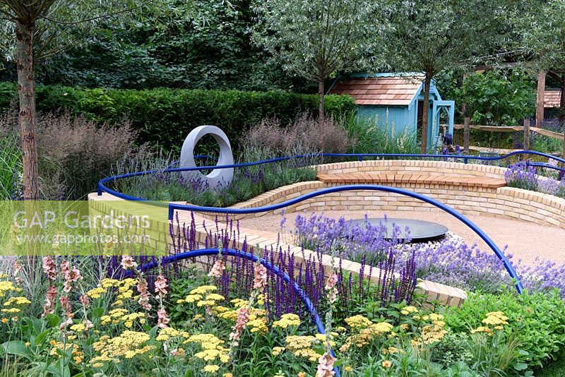 The Abbeyfield Society. A Breath Of Fresh Air. Patio area with central water bowl surrounded by curving seats with railings. External border with Achillea, salvia, verbascum and grasses. Designers: Rae Wilkinson. Sponsors: The Abbeyfield Society. RHS Hampton Court Palace Flower Show 2016