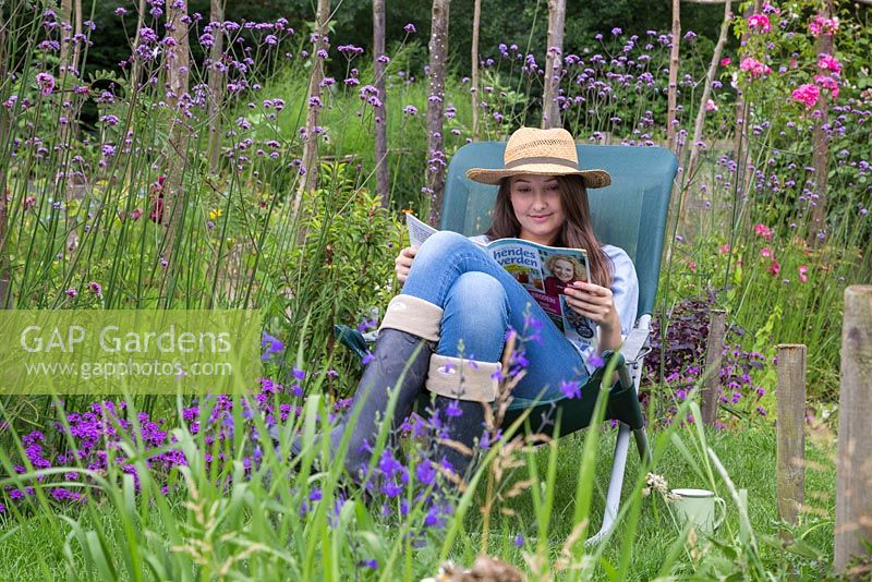 Girl sat reading a magazine in a deck chair beside a border of Verbena bonariensis