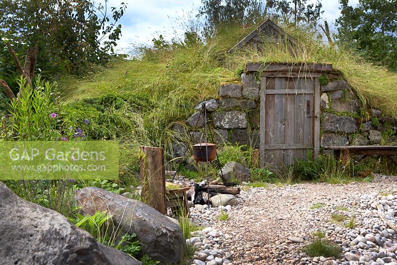 The Viking Cruises Scandinavian Garden. Basic house - dwelling in an earth mound covered by meadow grass and wildflowers. Fire pit with old seat and cooking pot. RHS Hampton Court Palace. Designer: Stephen Hall. Sponsor: Viking Cruises. 