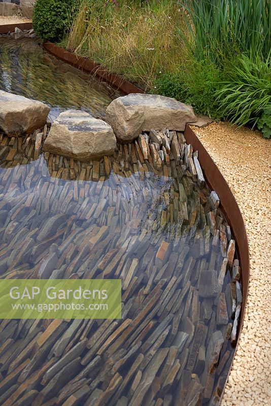 Stream with slate base layered at 90 degree angle. Steel edged with gravel path bordered by ferns and grasses. Zoflora. Outstanding Natural Beauty Garden, RHS Hampton Court Flower Show, 2016