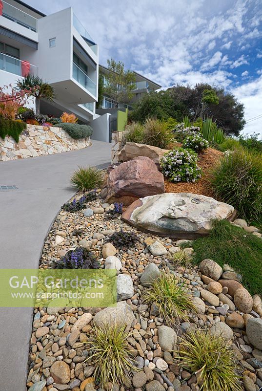 Driveway leading to a modern glass and concrete house with a steep sloping garden stabilised with large sandstone rocks, river pebbles and a variety of plants, shrubs and grasses. 