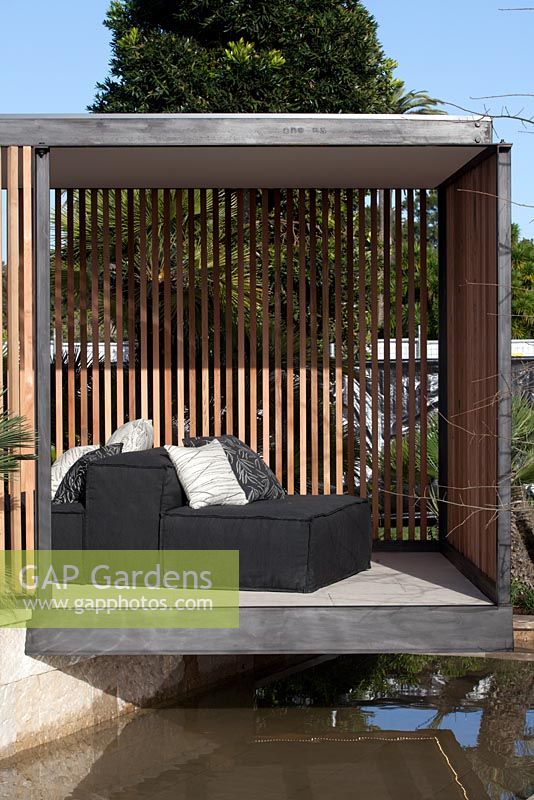 A black steel framed garden pavilion sits on top of a wall above a water feature, with lounge seats with cushions