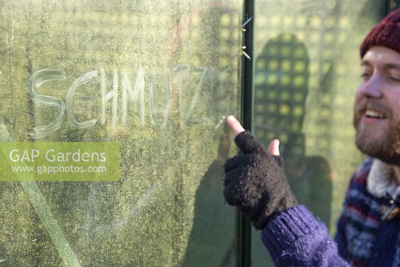 Mischievous man writing 'Schmutzig' on a dirty greenhouse glass pane