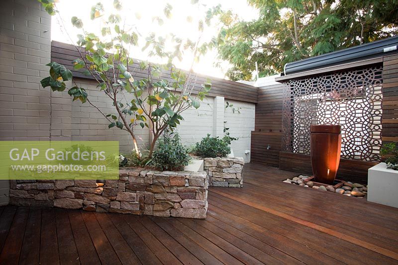 Inner city courtyard with featuring a raised bed with stone stack wall with a Cercis tree, a freestanding ceramic water feature, a rusty corten decorative screen and a timber slat panel wall


