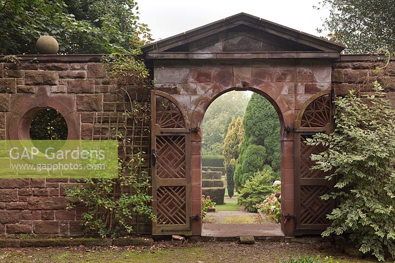 A view through a pseudo-classical style red sandstone lattice gateway to conifers in garden beyond - October, Burton Manor, Cheshire