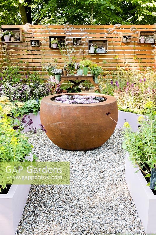 View of the garden with circular water feature, wooden work table with bouquets in pots and containers surrounded by wooden fence and raised bed with summer flowers including Achillea 'Terracotta', Alchemilla mollis, Astrantia 'Pink Pride', Euphorbia schillingii, Valeriana officinalis. Katie's Lymphoedema Fund: Katie's Garden. Designers: Carolyn Dunster, Noemi Mercurelli, RHS Hampton Court Palace Flower Show 2016 