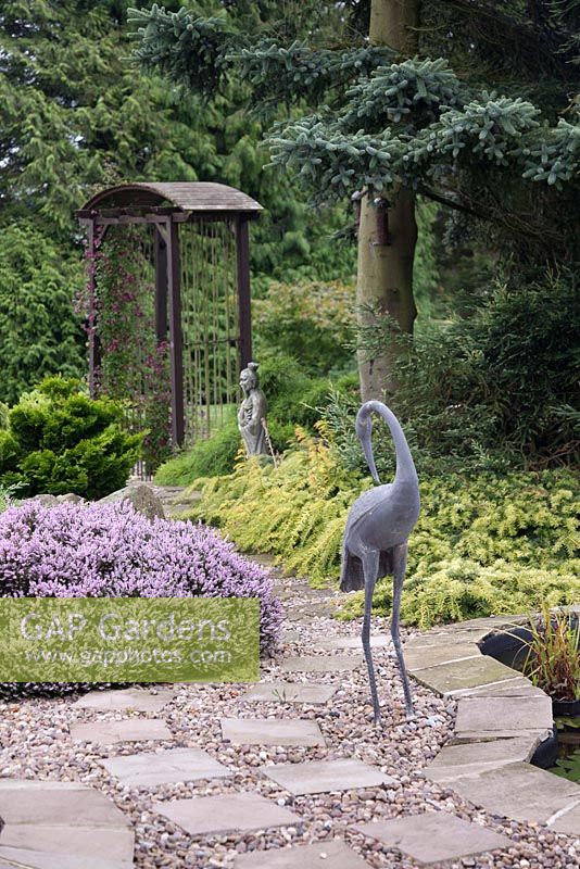 A metal heron sculpture at a pond edge with archway and trees beyond in a Japanese style garden.  June, North Yorkshire.