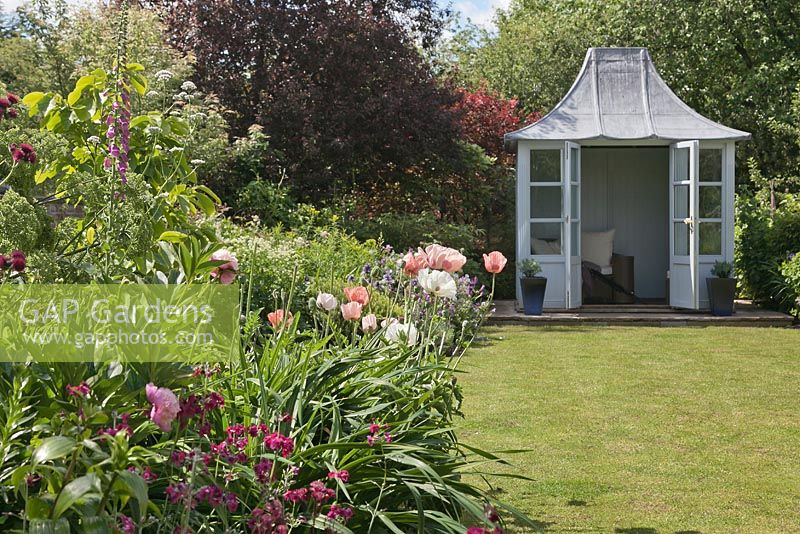 View across lawn to a pale blue painted summerhouse with seats and cushions in cottage garden with summer border. Cheshire, June. 