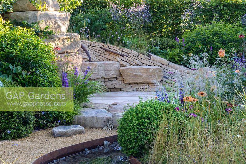 Dry stone seating area with steel tree root sculpture, herbaceous border in foreground with Cirsium rivulare, Echinacea and Campanula - Zoflora: Outstanding Natural Beauty, RHS Hampton Court Palace Flower Show 2016