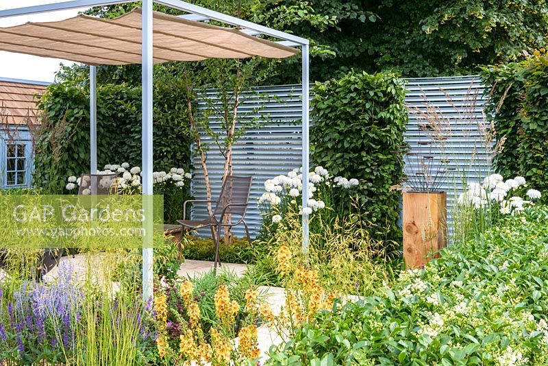 Steel pergola with canvas over a table and chairs on white paved raised terrace area, drought tolerant plants including verbascum, echinaceas, artemisia, geum, perovskia, stipa gigantea and salvias. Retreat Garden, RHS Hampton Court Flower Show in 2016