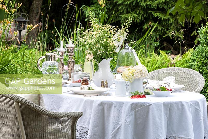A laid white table with silver candle holders, a bunch of flowers, glass jar and carafe and wicker chairs