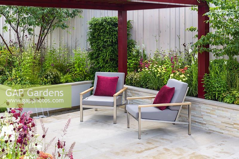 A pair of grey padded stainless steel Barlow Tyrie Equinox' Armchairs with burgundy cushions and pergola on Yorkstone paved terrace, surrounded by raised beds. Plants include Achillea millefolium 'Paprika', Pennisetum orientale, Penstemon 'Port Wine', Sanguisorba 'Chocolate Tip', Taxus baccata and Prunus serrula trees. Squire's 80th Anniversary Garden, RHS Hampton Court Flower Show 2016. Designer: Catherine MacDonald - Sponsor: Squire's Garden Centres