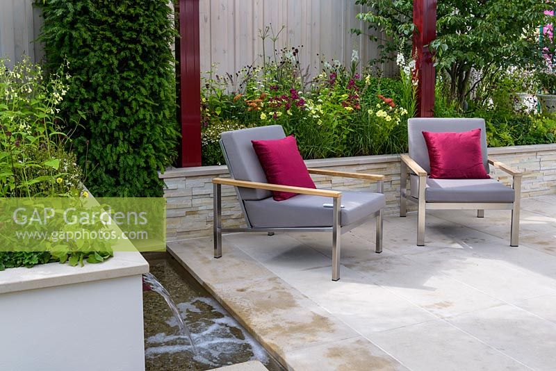 A pair of grey padded stainless steel Barlow Tyrie Equinox' Armchairs  with burgundy cushions and pergola on Yorkstone paved terrace, surrounded by raised beds. Plants include Achillea millefolium 'Paprika', Pennisetum orientale, Penstemon 'Port Wine', Sanguisorba 'Chocolate Tip' and Taxus baccata.  Squire's 80th Anniversary Garden, RHS Hampton Court Flower Show 2016. Designer: Catherine MacDonald - Sponsor: Squire's Garden Centres