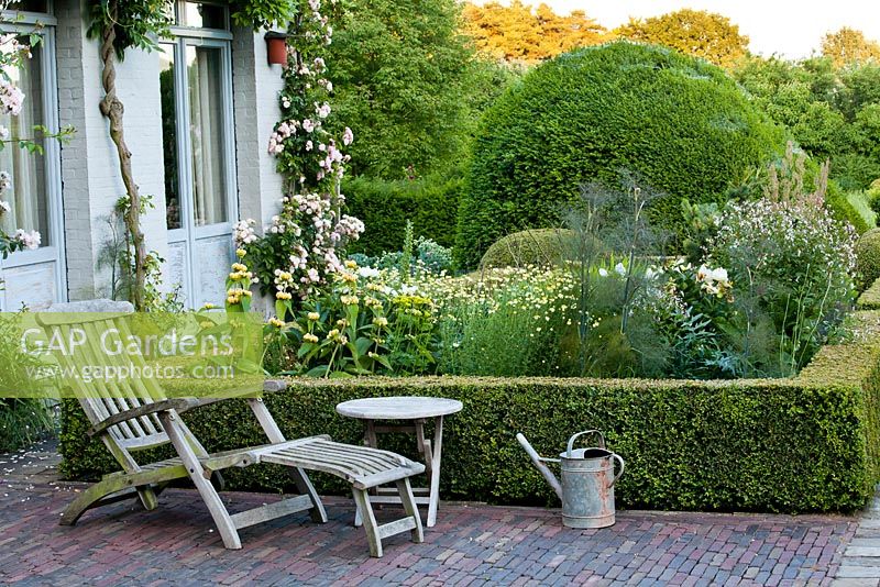 Lounger and table on a patio. Border of Gillenia trifoliata, Phlomis russeliana, fennel, Anthemis tinctoria, box topiary.