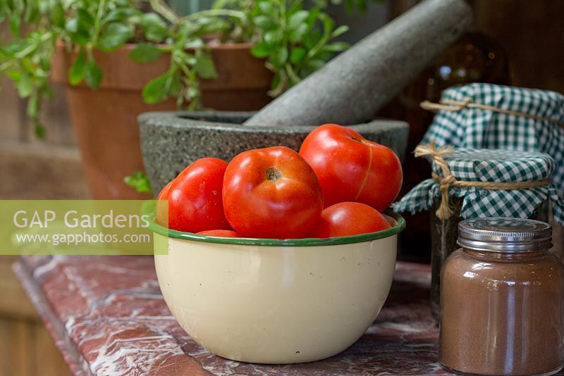 An old enamel bowl of Solanum lycopersicum, tomatoes on a stone counter top with jars of pickles and a stone mortar and pestle.