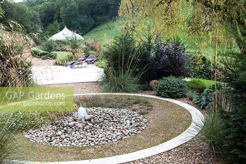 Circular water feature with water jet on pebbles and planting of Stipa gigantea Hebe and Taxus baccata