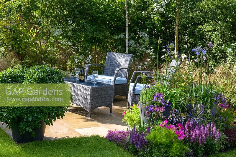Brick path leading to rectangular seating area with with synthetic wicker armchairs and table surrounded by mixed planting of Betula Jacquemontii, Anthriscus sylvestris, Cosmos,  Nepeta x faassenii 'Blue Wonder', Sambucus nigra porphyrophylla 'Eva' and pot with Pittosporum Tobira in A Garden by Association at RHS Hampton Court Flower Show 2015. Designer: Tina Vallis, MSGD