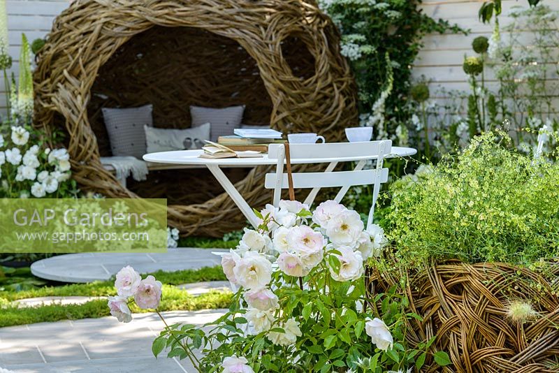 White metal bistro table and chairs and woven willow bird-hide with white and green plants including Rosa 'Iceberg - Living Landscapes 'City Twitchers' Garden, RHS Hampton Court Flower Show 2015. Designed by Sarah Keyser. CouCou Design