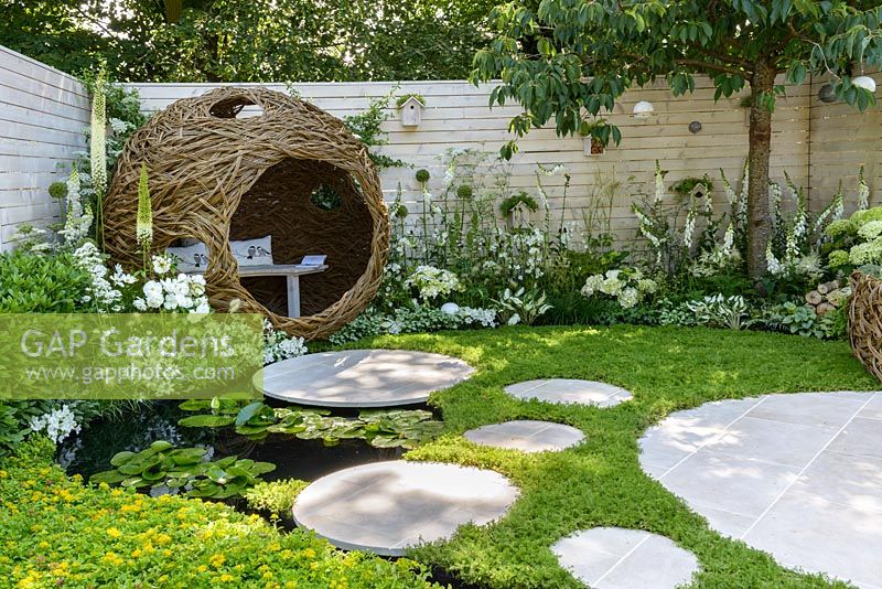 Wildlife-friendly small urban garden with spherical woven willow bird hide with bench, circular stepping stones, white flower borders with fencing, Chamomile nobile 'Treneague' lawn and pond with Nymphaea and rill. Living Landscapes 'City Twitchers' Garden, RHS Hampton Court Flower Show 2015. Designed by Sarah Keyser. CouCou Design