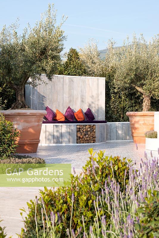 Outdoor seating with log storage, Olive trees in terracotta planters - The Retreat, RHS Malvern Spring Festival 2017 - Design: Villaggio Verde