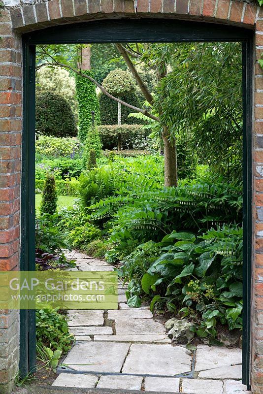 The entrance to a fifth-of-an-acre walled garden with stone path and shady border. Plants include Polygonatum x hybridum, Trachystemon orientalis, hosta, ferns and conical box.