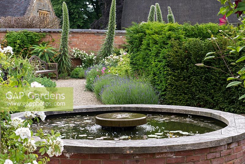 A walled garden with circular raised pond, Behind a path lined with lavender and roses leads to the dramatic flower spikes of Echium pininana.