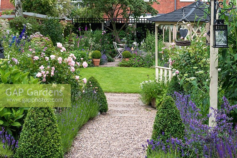 A 24m x 7.6m rear town garden. Lavender and box-edged pebble path leads past gazebo to circular lawn enclosed in cottage style herbaceous borders. At far end, there is shady seating, a compost bin amongst foxgloves, and work area.