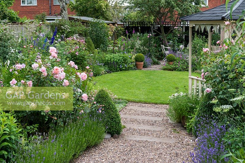 A lavender and box-edged pebble path leads past gazebo to circular lawn enclosed in cottage style herbaceous borders. At far end, there is shady seating, a compost bin amongst foxgloves, and work area.
