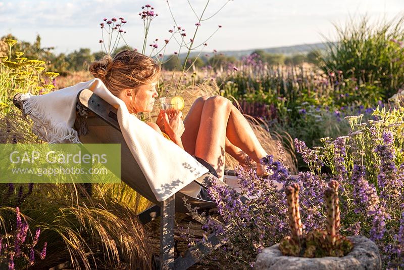 Young woman relaxing with a glass of lemonade in a deckchair on a boardwalk among naturalistic planting featuring perennials and grasses - July, Maison de l'arbre, France