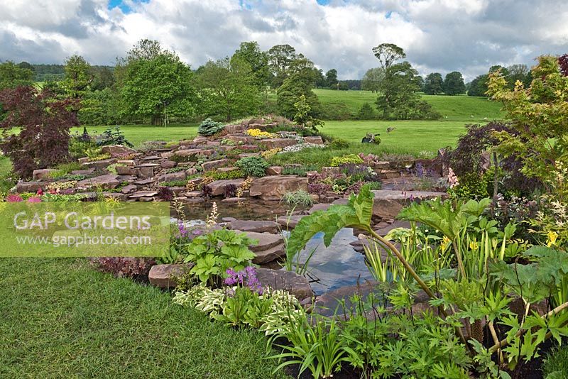 Jackie Knight's Just Add Water at RHS Chatsworth Flower Show 2017. Naturalistic water garden with pink sandstone rock, stepping stone path and waterside plants. Design: Jackie Sutton