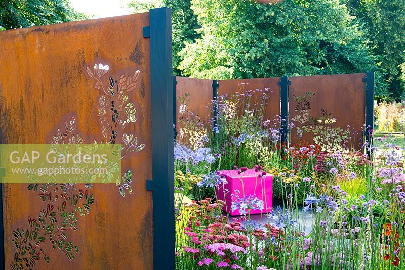 Decorative rusted steel panels enclose a colourful garden with pink cubed seating, plants include Verbena bonariensis, Cirsium rivulare 'Atropurpureum' and Agapanthus 'Blue Triumphator' - Colour Box, RHS Hampton Court Palace Flower Show 2017