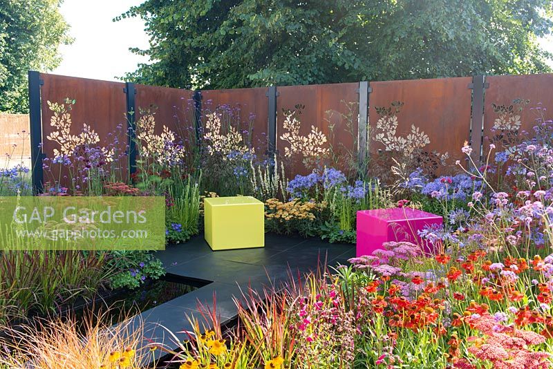 Decorative rusted steel panels enclose a colourful garden with pink and lime green cubed seating, plants include Helenium 'Moerheim Beauty', Penstemon 'Garnet', Agapanthus 'Blue Triumphator', Verbena bonariensis and Imperata cylindrica 'Rubra'- Colour Box, RHS Hampton Court Palace Flower Show 2017