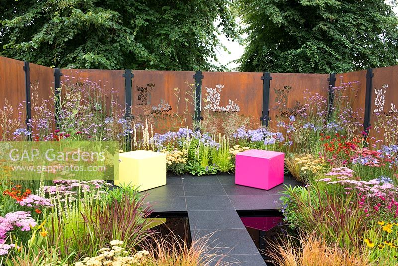 Decorative rusted steel panels enclose a colourful garden with pink and lime green cubed seating, plants include Helenium 'Moerheim Beauty', Penstemon 'Garnet', Agapanthus 'Blue Triumphator', Verbena bonariensis, Achillea 'Pretty Belinda', Achillea 'New Vintage Red' and Imperata cylindrica 'Rubra'- Colour Box, RHS Hampton Court Palace Flower Show 2017
