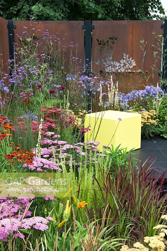 Decorative rusted steel panels enclose a colourful garden with lime green cubed seating, plants include, Agapanthus 'Blue Triumphator', Verbena bonariensis, Achillea 'New Vintage Red', Achillea 'Pretty Belinda', Verbascum chaixii 'Album', Imperata cylindrica 'Rubra' and Liatris spicata 'Alba' - Colour Box, RHS Hampton Court Palace Flower Show 2017