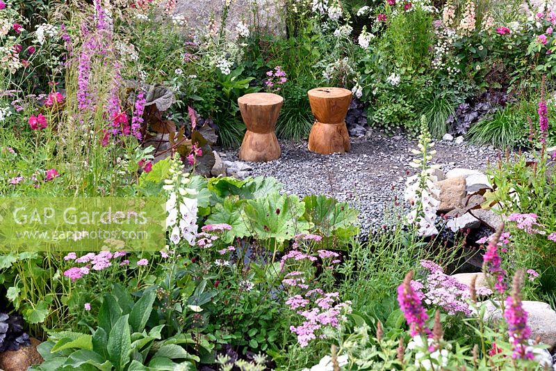 The Oregon Garden. Carved timber seats in gravel area surrounded by deep reds,cream and pink colour themed planting. Design: Sadie May Stuios. RHS Hampton Court Palace Flower Show 2017
