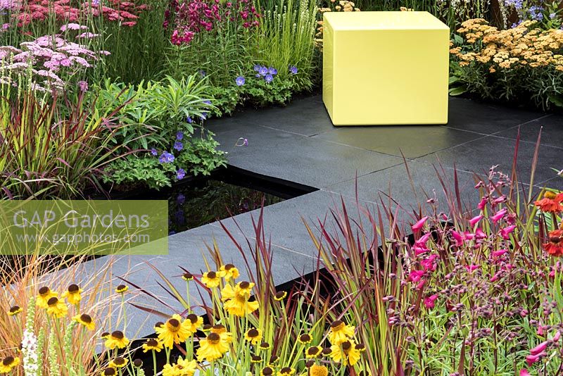 A small garden full of colourful perennials with  a pale yellow cube, two rectangular pools surrounded by slate paving. Perennials include: Achillea 'Taygatea' and 'Pink Island', Helenium 'Pipsqueak', Penstemon 'Garnet', Blood Grass - Imperata 'Red Baron' and Campanulas. Designed by Charlie Bloom.