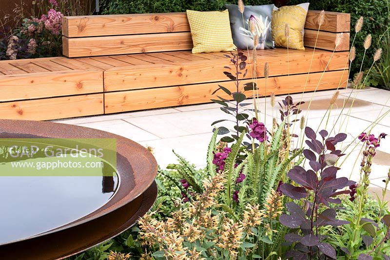 A small garden with circular steel bird bath and a long wooden bench is an ideal place to meet.  Plants include: Agastache 'Kudo Gold', Alchemilla mollis, Penstemon, Pennisetum thunbergii 'Red Buttons' and Hart's Tongue Ferns. Designed by: The Association of Professional Landscapers