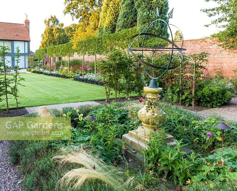 An armillary sphere provides a focal point in a modern Cheshire country garden, designed by Louise Harrison-Holland. Planting includes roses and Stipa tenuissima. Beyond, plants include pleached Pyrus calleryana 'Chanticleer', Japanese anemones, Stachys, Persicaria, Ophiopogon planiscapus 'Nigrescens' and Miscanthus sinensis 'Kleine Fontaine'