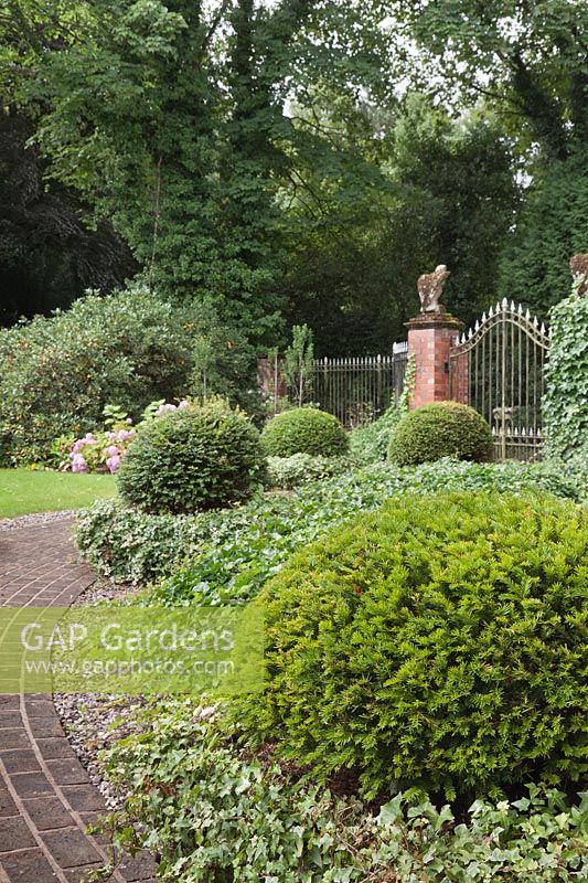 A red brick path leading to classical black iron railings with stone falcons and yew topiary balls on living plinths of ivy