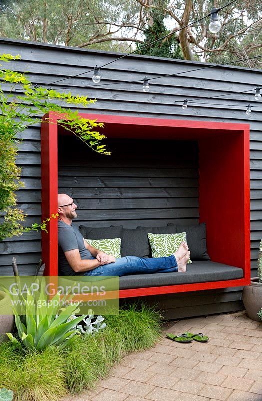 Garden owner and designer Steven Wells, relaxing in a red painted timber sitting pod he built, attached to a black painted timber wall.