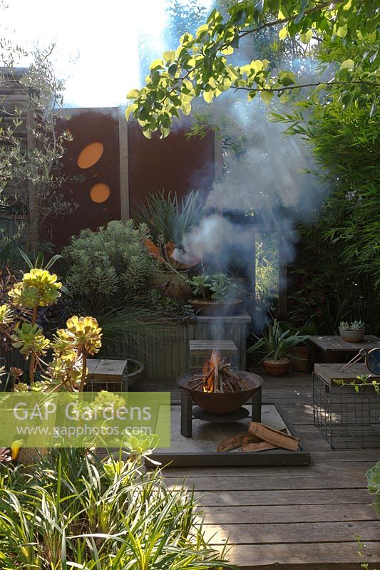 A sunken fire pit area with a freestanding metal fire pit, on a timber deck surrounded by raised garden beds and rusty coreten steel screens.